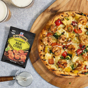 A grilled pizza topped with shrimp and pineapple sits on a wooden board next to a packet of McCormick Baja Citrus seasoning.