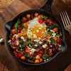 A skillet dish featuring a fried egg nestled among chickpeas, spinach, red peppers, and feta cheese, presented on a wooden board.