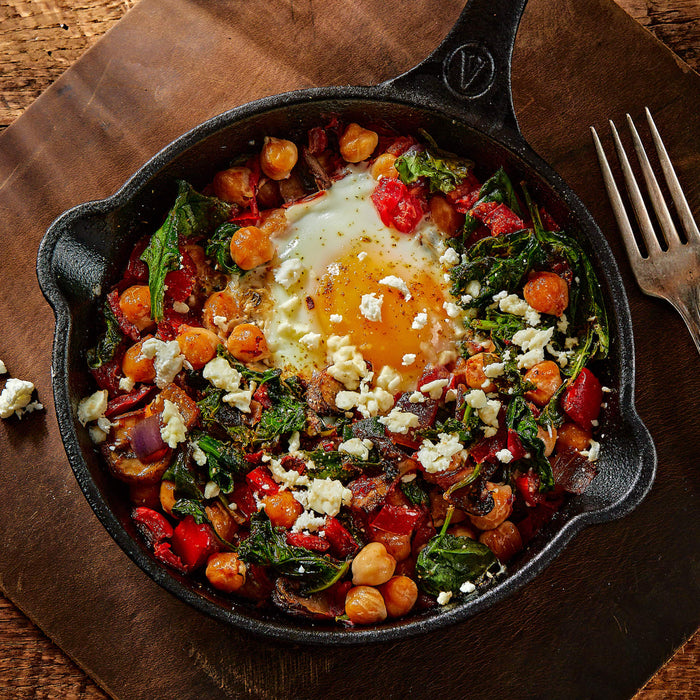 A skillet dish featuring a fried egg nestled among chickpeas, spinach, red peppers, and feta cheese, presented on a wooden board.