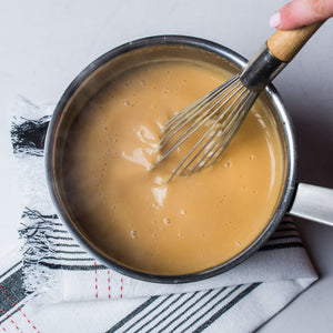 A whisk is stirring a creamy light brown sauce in a stainless steel saucepan.