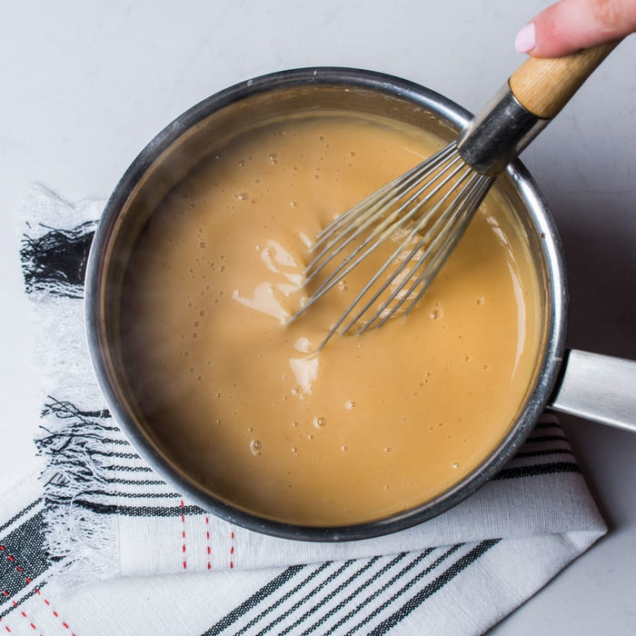 A whisk is stirring a creamy light brown sauce in a stainless steel saucepan.