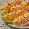 Four grilled corn on the cob topped with cheese and chili powder on a white plate with lime wedges.