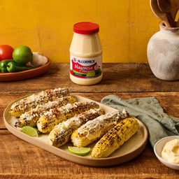 A plate of grilled corn on the cob topped with McCormick Mayonesa lime wedges and cotija cheese.