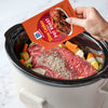 A person is adding a McCormick Slow Cookers Pot Roast seasoning packet to a slow cooker with a beef roast and vegetables.