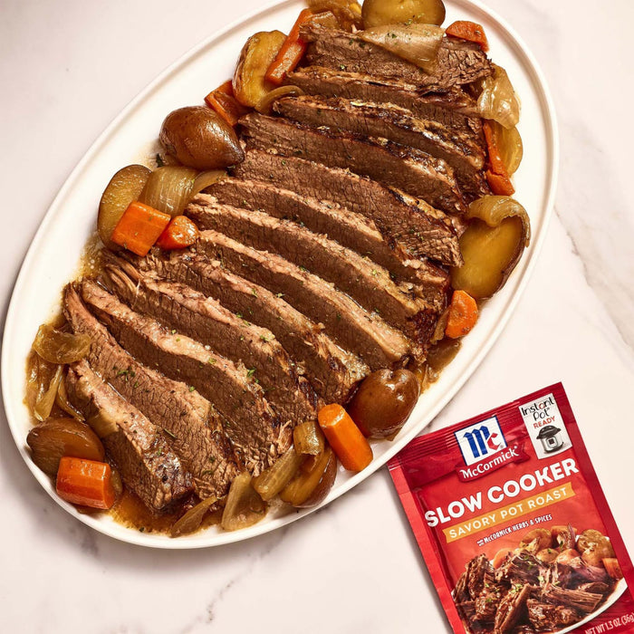 A plate of sliced pot roast with carrots, potatoes, and onions next to a McCormick Slow Cooker Savory Pot Roast seasoning packet.