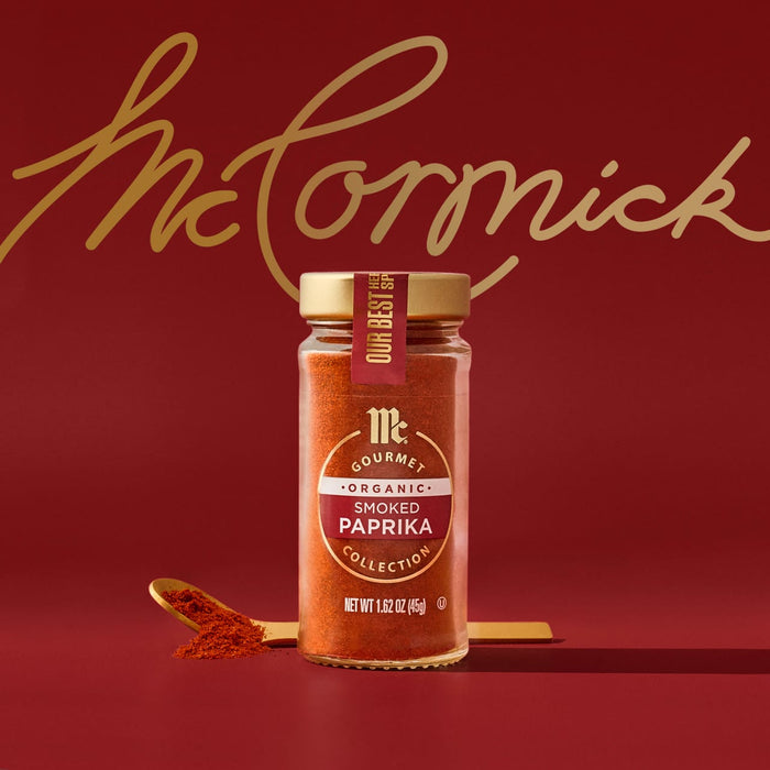 A glass jar of Mccormick Gourmet Smoked Paprika Organic 1.62 Oz sits on a red surface with a golden spoon holding some of the red spice, against a red background with the gold McCormick script logo.