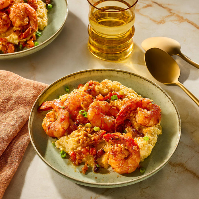 A plate of shrimp and grits, a Southern comfort food dish, is presented with a glass of beverage and a golden spoon.