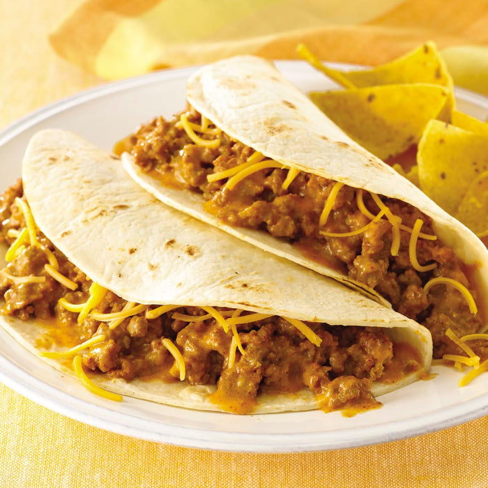 Two soft tacos filled with ground beef cheese and a creamy sauce are on a white plate with tortilla chips.