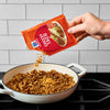 A hand pours McCormick Great Value Taco seasoning into a pan of cooked ground beef.