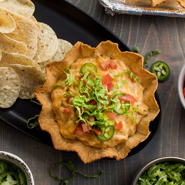 A delicious-looking taco dip in a tortilla bowl, garnished with lettuce, tomatoes, and jalapeños, served with tortilla chips.