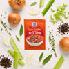 A packet of McCormick Slow Cookers Hearty Beef Stew seasoning mix is surrounded by fresh ingredients like onions, celery, and herbs.