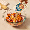 A hand pours a packet of Mccormick Toasted Onion And Garlic Seasoning Mix 1.25 Oz over a glass bowl of chopped root vegetables, including potatoes, carrots, and red onions.