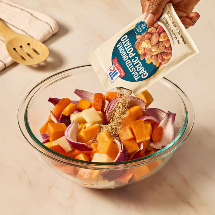 A hand pours a packet of Mccormick Toasted Onion And Garlic Seasoning Mix 1.25 Oz over a glass bowl of chopped root vegetables, including potatoes, carrots, and red onions.