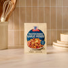 A packet of Mccormick Toasted Onion And Garlic Seasoning Mix 1.25 Oz is displayed on a kitchen counter, with kitchen utensils and plates visible in the background to suggest a home-cooked meal.