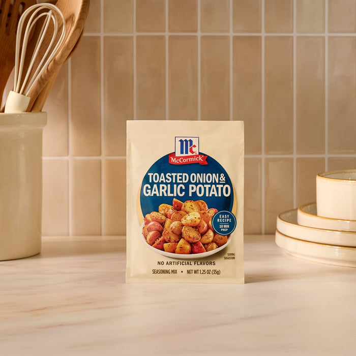 A packet of Mccormick Toasted Onion And Garlic Seasoning Mix 1.25 Oz is displayed on a kitchen counter, with kitchen utensils and plates visible in the background to suggest a home-cooked meal.