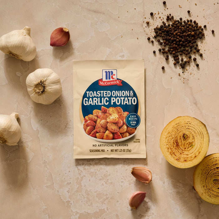 A packet of Mccormick Toasted Onion And Garlic Seasoning Mix 1.25 Oz is arranged on a countertop with fresh ingredients like garlic bulbs, a sliced onion, and black peppercorns.