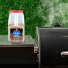 A container of Pit Boss Competition Blend wood pellets sits next to a smoker grill.