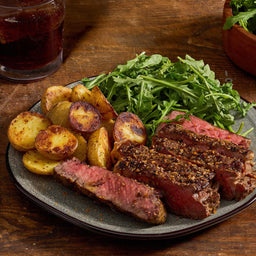 A plate of sliced steak seasoned with Mccormick Grill Mates Montreal Steak Seasoning, roasted potatoes, and a side of fresh arugula is presented on a wooden table next to a glass of iced tea.