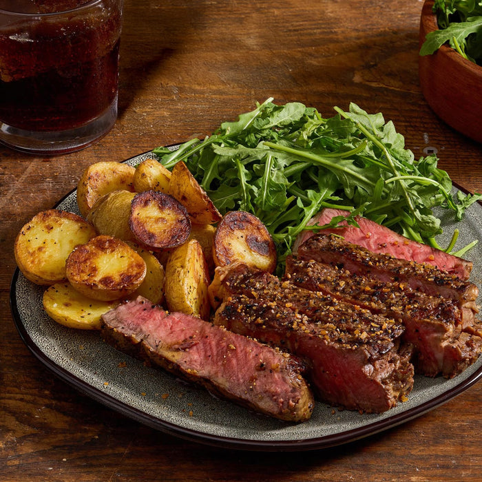 A plate of sliced steak seasoned with Mccormick Grill Mates Montreal Steak Seasoning, roasted potatoes, and a side of fresh arugula is presented on a wooden table next to a glass of iced tea.