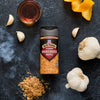 A bottle of Mccormick Grill Mates Smokehouse Maple Blend seasoning is centered on a dark surface, surrounded by garlic cloves, whole garlic bulbs, orange peels, a bowl of syrup, and a pile of brown sugar, with wisps of smoke on the left.