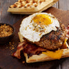 A waffle sandwich with bacon, cheese, a burger patty, and a fried egg on top sits on a wooden board next to a small bowl of seasoning.