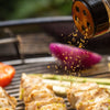 Seasoning is being sprinkled over chicken and vegetables on a grill.