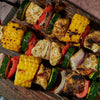 Three skewers of grilled chicken and vegetables including corn zucchini red pepper and onion are arranged on a wooden board.