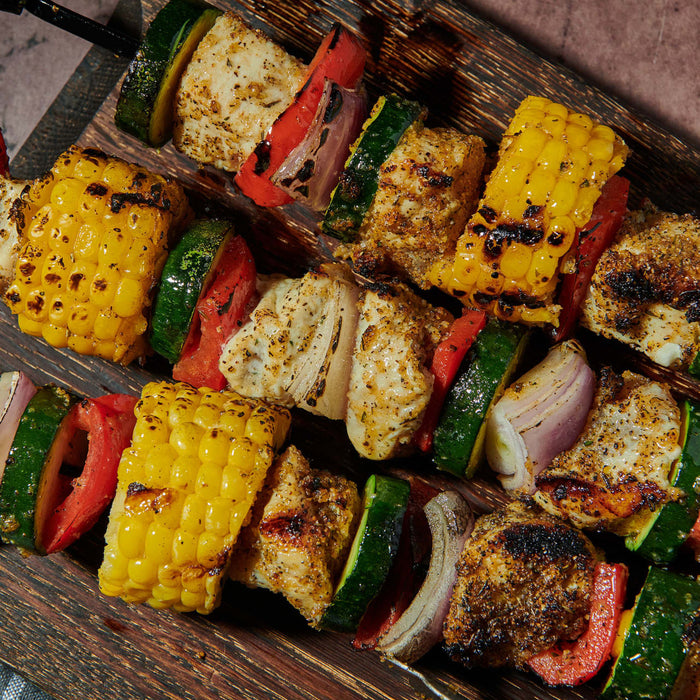 Three skewers of grilled chicken and vegetables including corn zucchini red pepper and onion are arranged on a wooden board.