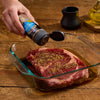 A person seasons a raw steak in a glass dish with McCormick Grill Mates Montreal Steak Seasoning.
