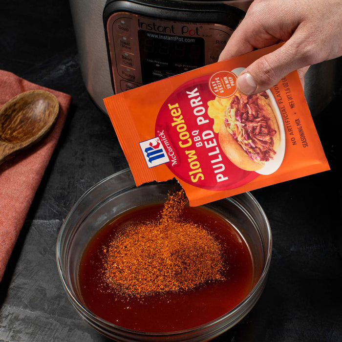 A hand pours McCormick Slow Cookers Pulled Pork seasoning into a bowl of sauce, preparing a meal with a slow cooker in the background.