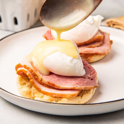 A plate of eggs benedict with hollandaise sauce being poured over the top.