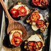 Grilled chicken breasts topped with melted mozzarella cheese and grilled tomato slices are arranged on a metal baking sheet with a wooden spatula.