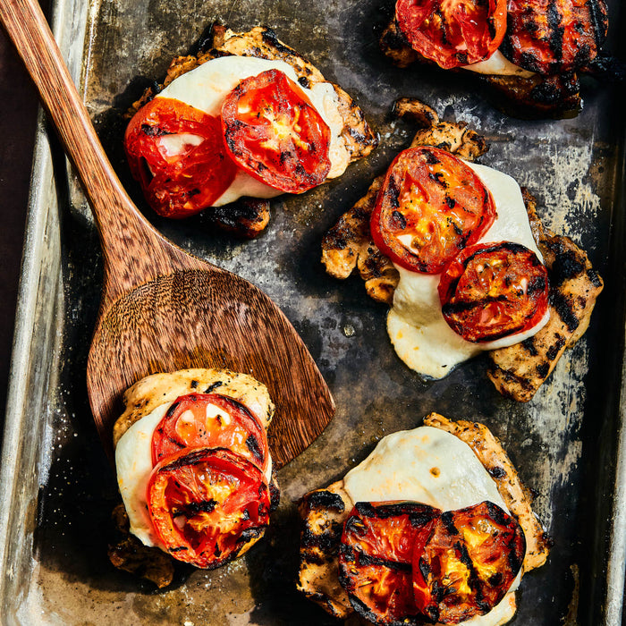 Grilled chicken breasts topped with melted mozzarella cheese and grilled tomato slices are arranged on a metal baking sheet with a wooden spatula.