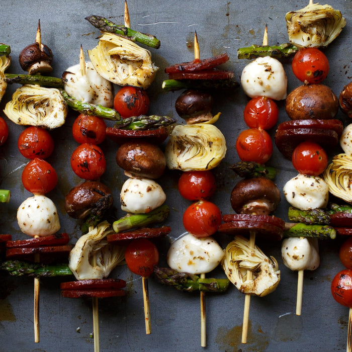 Grilled skewers with artichoke hearts tomatoes mozzarella mushrooms asparagus and sausage are arranged on a metal surface.