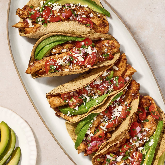 Four chicken tacos with avocado tomato and cheese on a white plate.