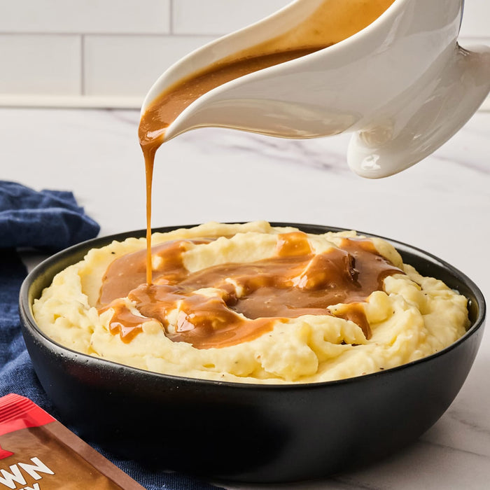 A bowl of mashed potatoes with brown gravy being poured from a white gravy boat, showcasing a comforting and classic dish.