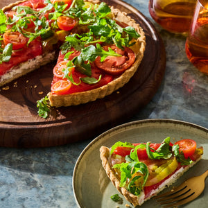 A colorful tomato tart topped with fresh greens is displayed on a wooden board, with a slice served on a plate.