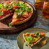A colorful tomato tart topped with fresh greens sits on a wooden board next to a slice on a plate, suggesting a fresh and appetizing dish.