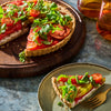 A vibrant tomato tart topped with fresh arugula is displayed on a wooden board and a plate, showcasing its colorful layers and inviting presentation.