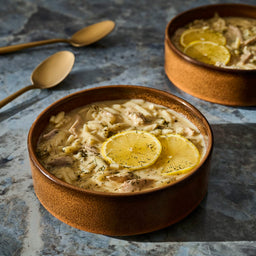 Two bowls of creamy lemon chicken soup are presented with golden spoons on a textured surface.