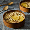 Two bowls of creamy chicken and orzo soup garnished with lemon slices are presented with golden spoons on a textured surface.