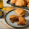 A close-up shot of golden-brown Jamaican patties, one broken open to reveal a savory filling, served on a blue plate with a side of green sauce.