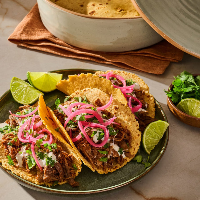 A vibrant plate of tacos topped with meat, pickled onions, cilantro, and lime wedges, alongside a bowl of tortillas, showcasing a delicious and inviting meal.