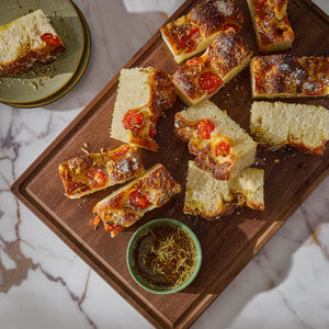 A wooden board displays slices of focaccia bread topped with tomatoes, alongside a small bowl of pesto, set against a marble background.