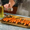 A person seasons baby carrots on a baking sheet with olive oil nearby, ready for roasting.