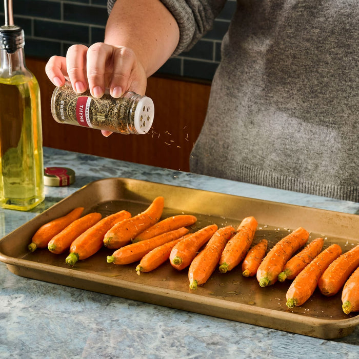 A person seasons baby carrots on a baking sheet with olive oil nearby, ready for roasting.