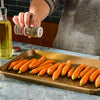 A person seasons baby carrots on a baking sheet with olive oil nearby, ready for roasting.