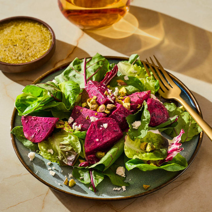 A vibrant salad featuring magenta beets, green lettuce, pistachios, and feta cheese, served on a blue plate with a side of dressing.