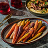 A plate of colorful roasted carrots sits alongside a bowl of braised meat over polenta and two glasses of red wine, suggesting a gourmet meal.