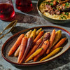 A plate of colorful roasted carrots sits alongside a bowl of braised meat over polenta and two glasses of red wine, suggesting a gourmet meal.